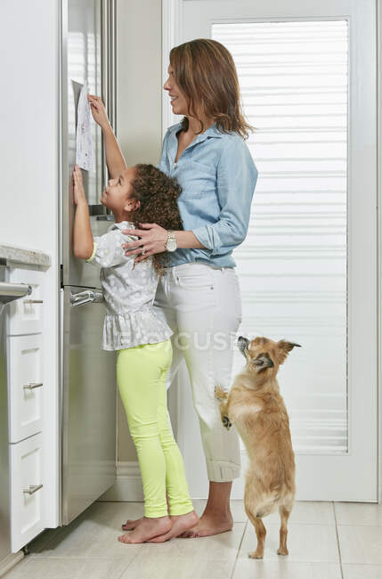 Side view of mother and daughter in kitchen sticking picture to fridge — Stock Photo