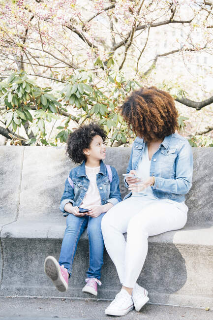 Madre e hija sentadas lado a lado sosteniendo teléfonos inteligentes, cara a cara - foto de stock