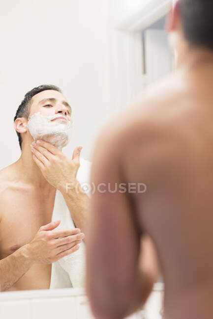 Hombre adulto medio, mirando en el espejo, aplicando espuma de afeitar al cuello, vista trasera - foto de stock