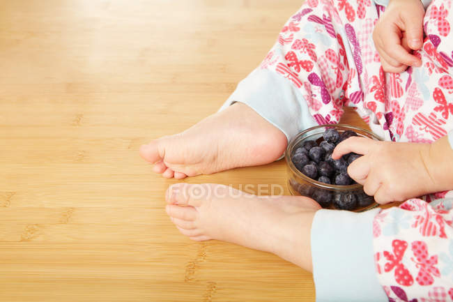 Cropped image of infant sitting on floor with blueberries in bowl — Stock Photo