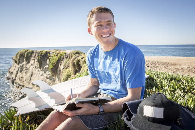 Young man sitting by cliff edge, writing in book, smiling — Stock Photo