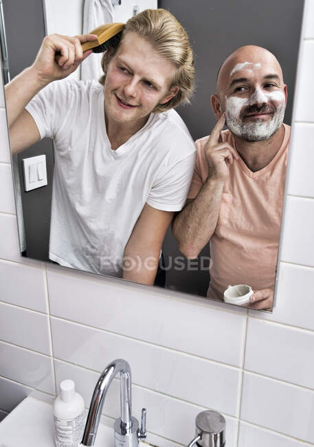 Bathroom mirror image of male couple shaving and brushing hair — Stock Photo