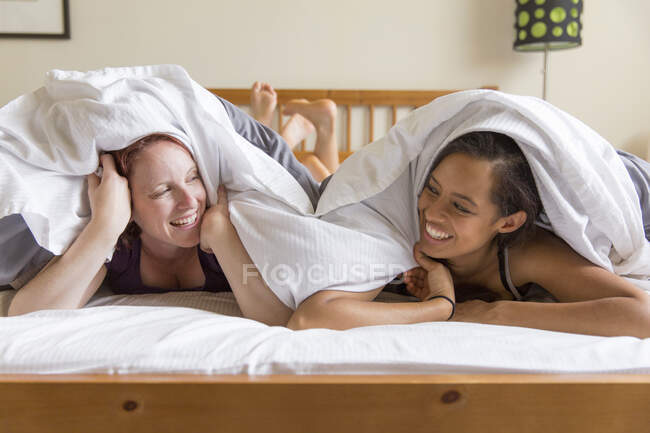 Young women lying on front in bed underneath quilt face to face smiling — Stock Photo