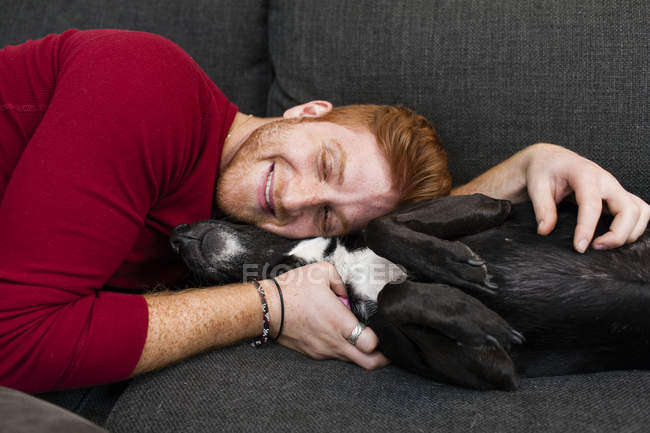 Jovem deitado no sofá abraçando animais de estimação olhos de cão fechado sorrindo — Fotografia de Stock
