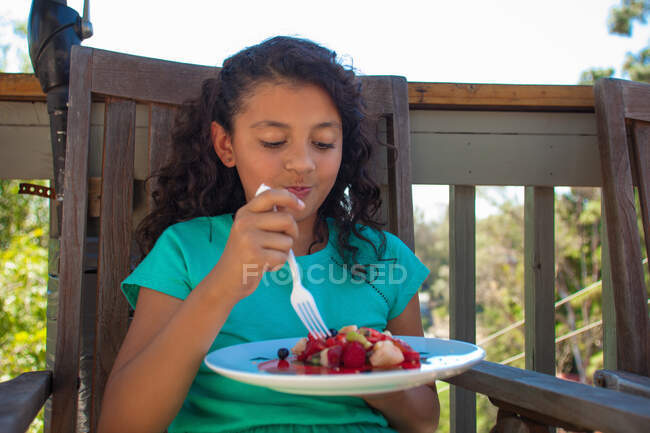 Chica comiendo postre de frutas en el banco del jardín - foto de stock