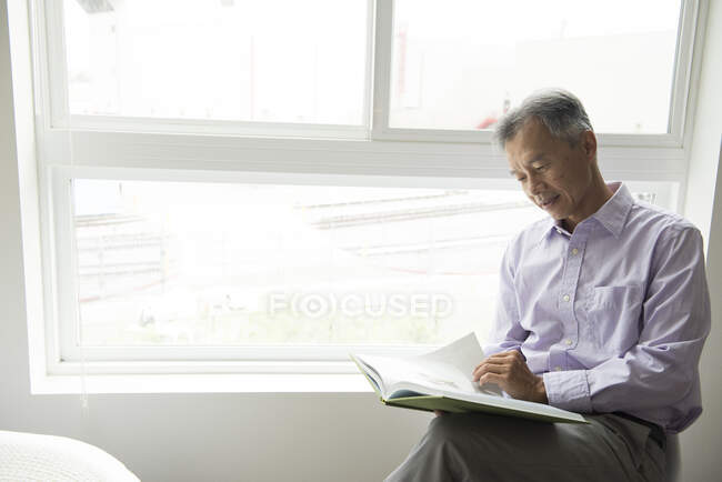 Maduro homem sentado na frente de janela leitura livro — Fotografia de Stock