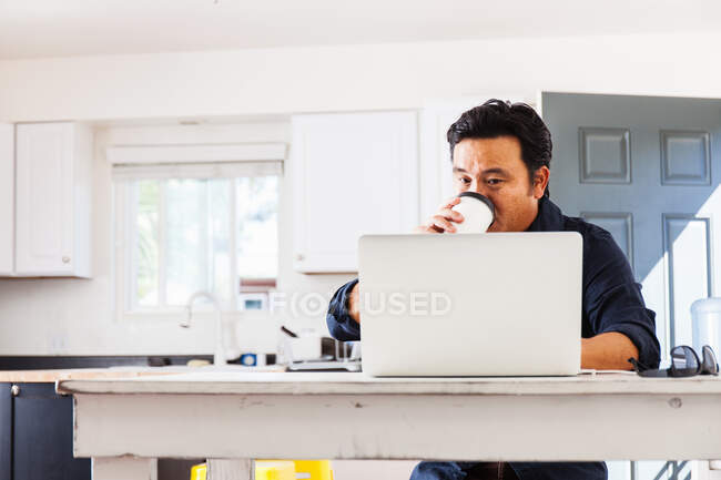 Reifer Geschäftsmann tippt am Laptop, während er am Küchentisch Kaffee zum Mitnehmen trinkt — Stockfoto