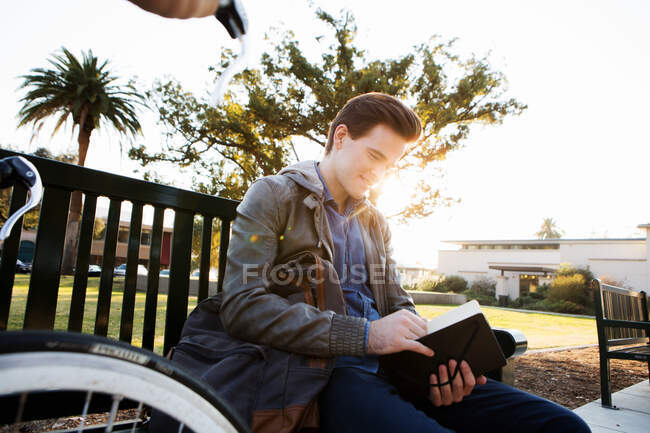 Jovem homem lendo livro no banco do parque iluminado pelo sol — Fotografia de Stock