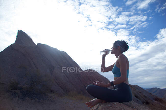Женщина, сидящая со скрещенными ногами у скал Васкез, пьет бутылку воды — стоковое фото