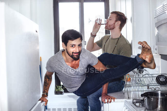 Мужская пара дома, мужчина среднего возраста пьет, пока его партнер балансирует на кухонной технике — стоковое фото