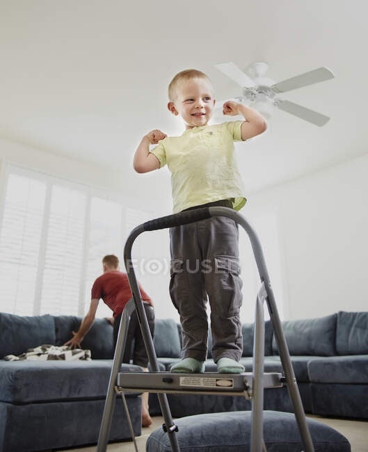 Menino flexão muscular na sala de estar, pai no fundo — Fotografia de Stock