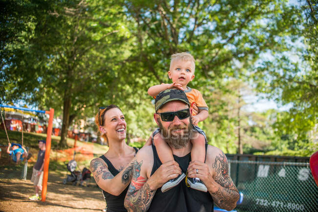 Tatuado padre llevando bebé niño en hombros mirando a la cámara sonriendo - foto de stock