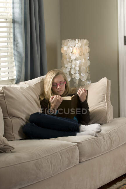 Pre adolescente chica reclinado en sofá lectura libro - foto de stock