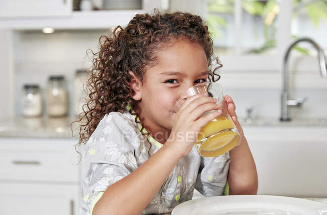 Chica en el mostrador de cocina bebiendo jugo de naranja mirando a la cámara - foto de stock