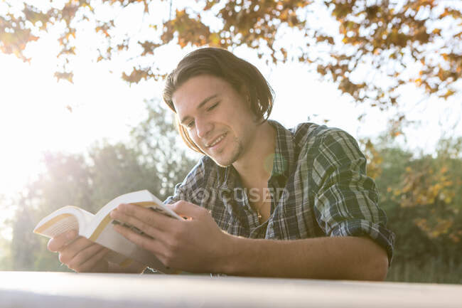 Jeune homme dehors assis à la table regardant vers le bas le livre de lecture, souriant — Photo de stock
