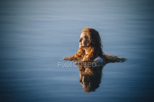 Head and shoulders of young woman with long red hair in lake with eyes closed — Stock Photo