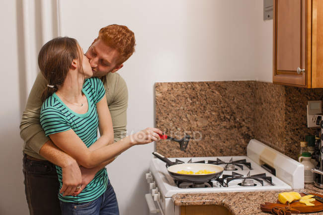 Молодой человек на кухне обнимает молодую женщину, готовит на плите, целует — стоковое фото