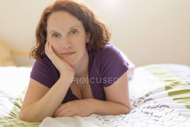 Giovane donna sdraiata sul davanti sul letto, mano sul mento guardando la fotocamera — Foto stock
