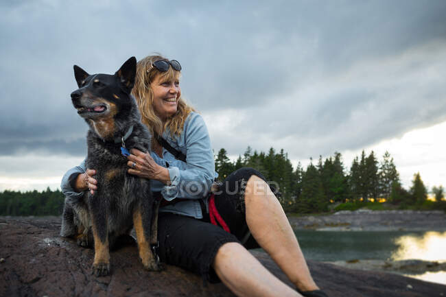 Mujer mayor sentada en roca abrazando perro en la costa de Maine, EE.UU. - foto de stock
