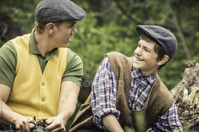 Man and boy wearing flat caps whittling twig with penknife, face to face smiling — Stock Photo