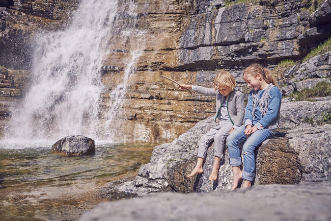 Hermano y hermana, sentados en la roca, relajados, junto a la cascada - foto de stock