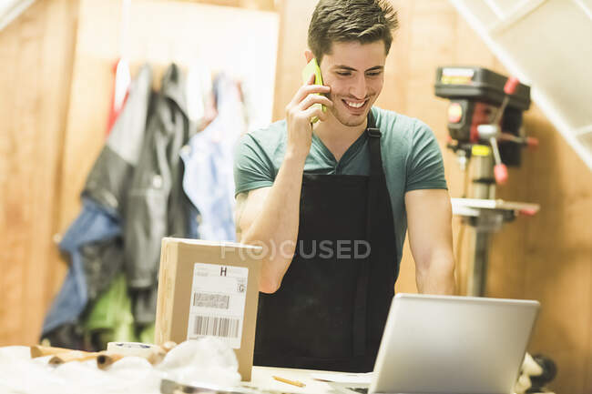 Молодий чоловік у майстерні стоїть за столом, розмовляючи по телефону, дивлячись на ноутбук — стокове фото