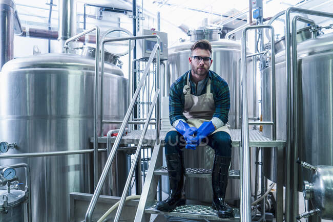 Young man in brewery wearing apron, protective gloves and wellington boots sitting on steps looking at camera — Stock Photo
