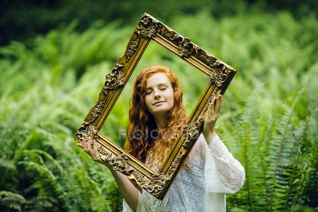 Giovane donna che tiene cornice d'oro tra felci della foresta — Foto stock