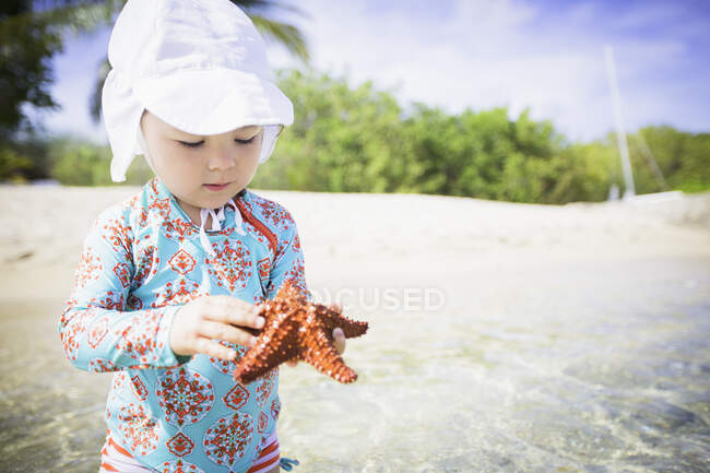 Menina na praia vestindo roupa de banho e chapéu de sol segurando estrela do mar olhando para baixo, St. Croix, Ilhas Virgens dos EUA — Fotografia de Stock