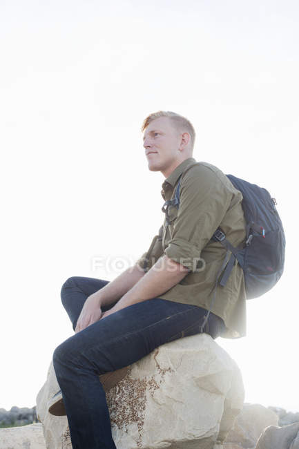 Giovane uomo che indossa lo zaino seduto sulla roccia guardando altrove — Foto stock