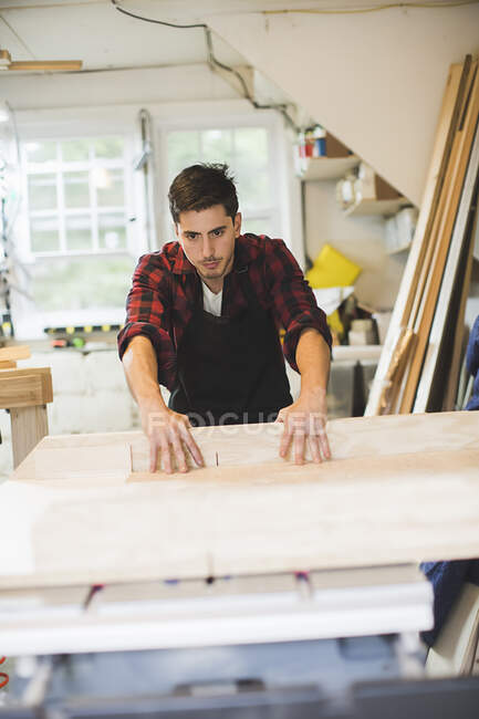 Jovem em oficina vestindo avental usando serra de mesa para cortar madeira — Fotografia de Stock