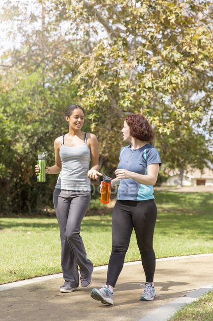 Giovani donne fuori a piedi indossando abbigliamento sportivo che trasportano bottiglie d'acqua parlando — Foto stock