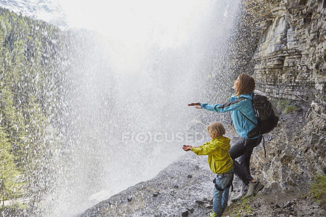 Mutter und Sohn stehen unter dem Wasserfall, reichen sich die Hände, um das Wasser zu spüren, Rückansicht — Stockfoto