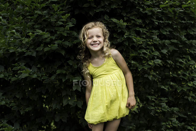 Портрет посміхаючись світле волосся дівчини носити жовте плаття стоячи перед саду хедж — стокове фото