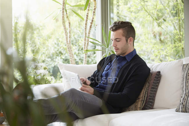 Чоловік розслабляється на дивані з ногами вгору дивиться на цифровий планшет — стокове фото