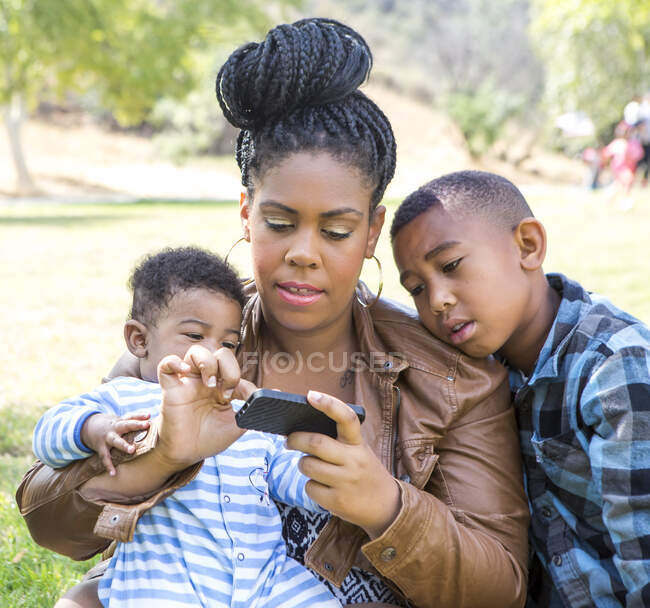 Madre e hijos sentados mensajes de texto en el teléfono inteligente mirando hacia abajo - foto de stock