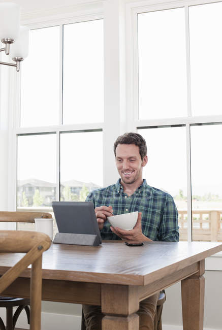 Mittlerer erwachsener Mann frühstückt, während er ein digitales Tablet betrachtet — Stockfoto