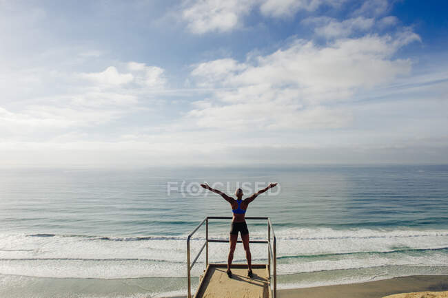 Junge Frau steht auf Aussichtspunkt am Meer, Arme erhoben, Rückansicht — Stockfoto