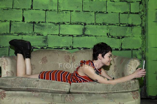 Середня доросла жінка розслабляється на зношеному дивані, використовуючи смартфон — стокове фото