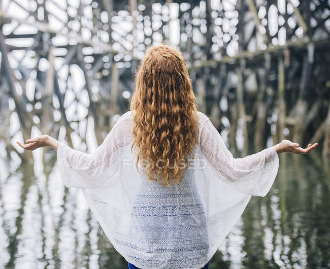 Rear view of woman with long red hair meditating in front of wood river pier — Stock Photo