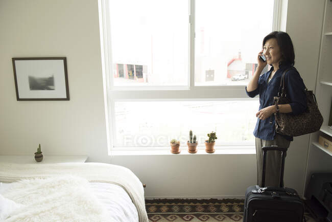 Mujer madura de pie en la habitación del hotel con la maleta usando el teléfono inteligente - foto de stock