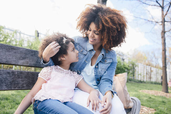 Madre con el brazo alrededor de la hija sentada en el banco del parque cara a cara - foto de stock