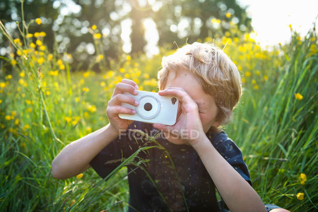 Niño sentado en la hierba larga tomar fotografías con el teléfono inteligente - foto de stock