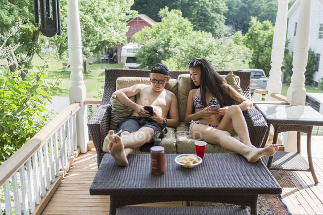 Подростковая пара в купальниках на крыльце ест закуски и смотрит на смартфоны — стоковое фото
