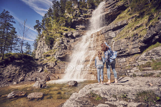Мать и дочь стоят на скалах, наблюдая водопад, вид сзади — стоковое фото