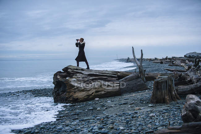 Woman looking through binoculars from large driftwood tree stump on beach — Stock Photo