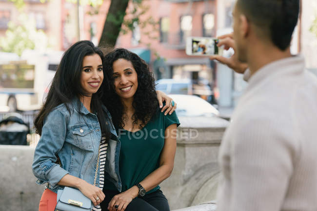 Mid adult man taking smartphone photograph of female friends in city park — Stock Photo