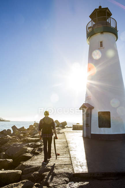 Young man holding skateboard, walking beside lighthouse, rear view — Stock Photo