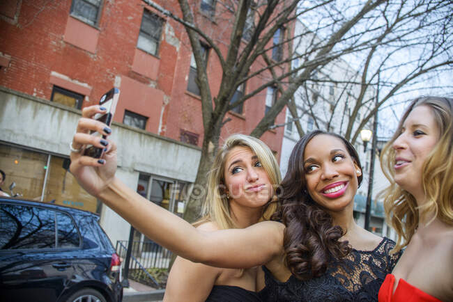 Three glamorous young adult female friends taking smartphone selfie on street — Stock Photo