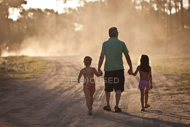 Padre e hijas caminando de la mano en el camino del polvo - foto de stock
